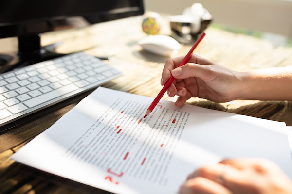 Close-up Of A Person's Hand Marking Errors With Red Colored Pencil On A Piece Of Paper