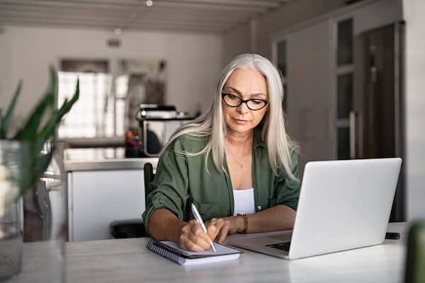 an image of an older woman with glasses wearing a green shirt writing in a notepad with a pen in front of her opened laptop