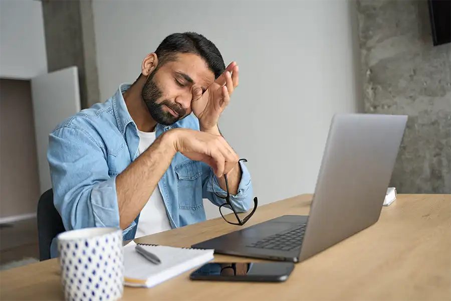 exhausted author rubbing his eyes while sitting in their modern home office with a laptop, notebook and cup of coffee on a wooden table