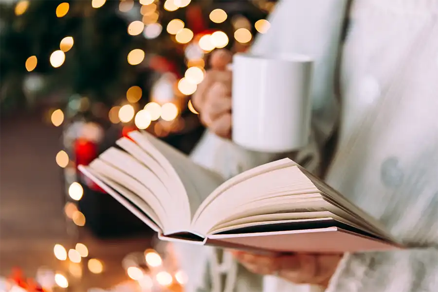 close up image of a woman's hands. One is holding an open book and her other hand is holding a white coffee mug. There are Christmas lights behind her, but they are out of focus