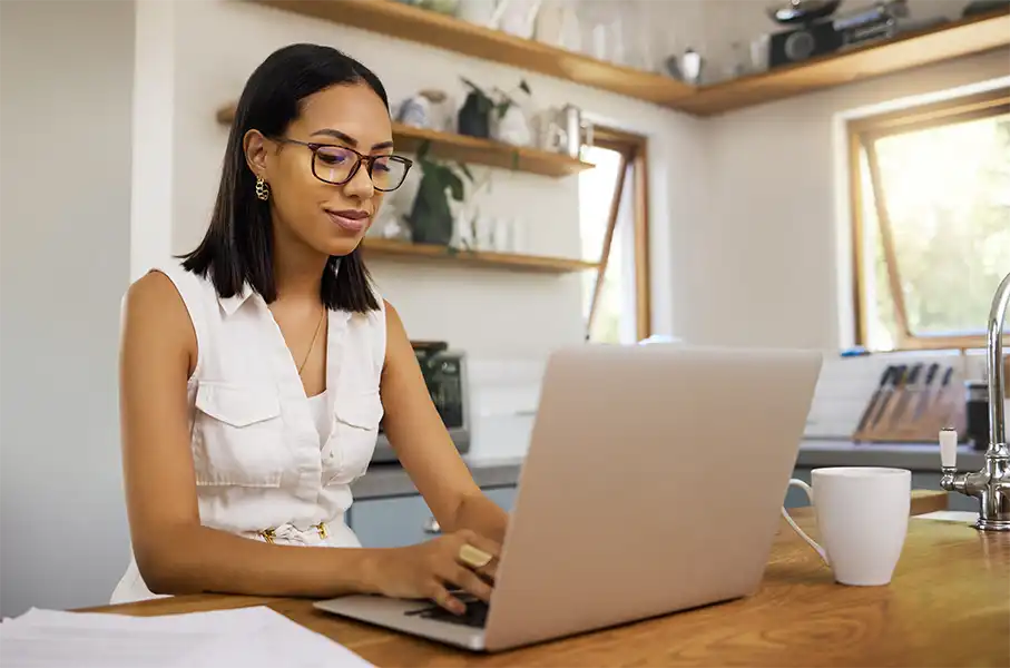 a women with black chic hair wearing all white typing on a laptop in her kitchen. She has notes and a white coffee cup next to her laptop.