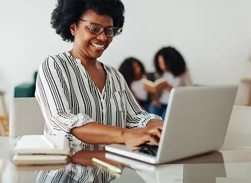 an african american author typing on her laptop in her living room at a glass table. you can see two kids reading a book together in the background sitting on a couch