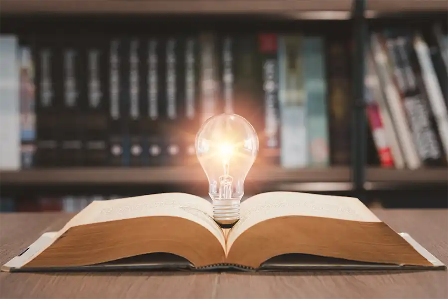 An open book resting on a wooden table in front of a large bookshelf. the book has a lightbulb coming out of the middle of the book.