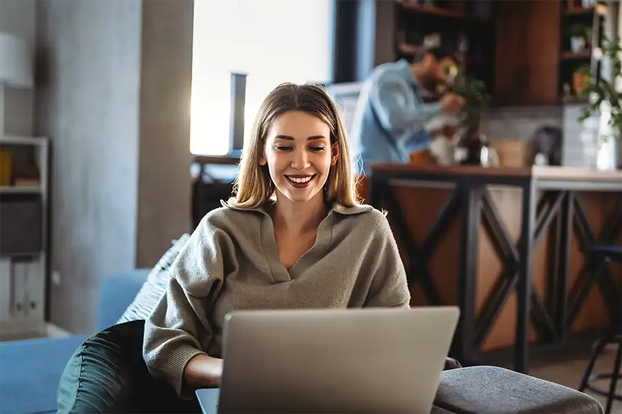 A woman in a gray sweater and maroon lipstick is sitting in her living room working on her laptop