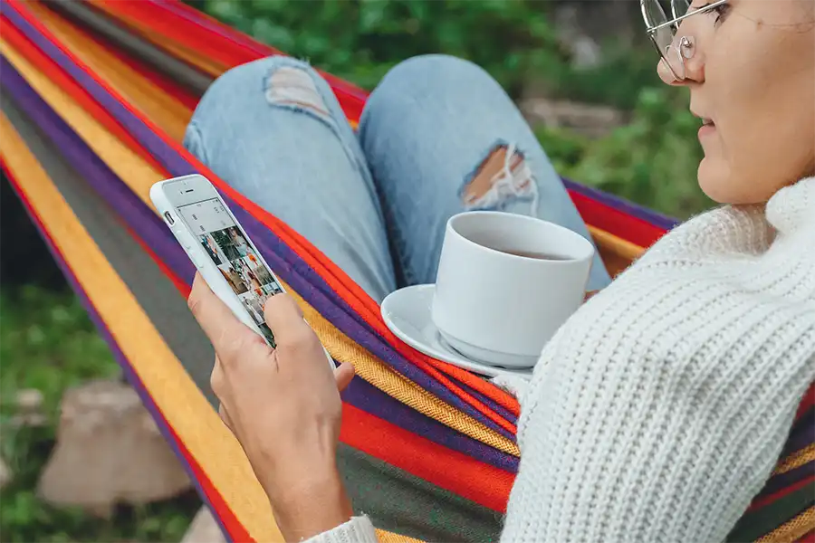 Young woman laying in a hammock while drinking coffee and using her Iphone to go on Instagram