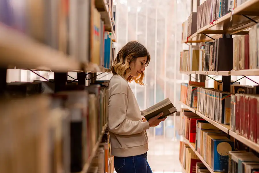 Young woman with a curly bob, gray sweater and jeans standing in an aisle in a library reading a book.