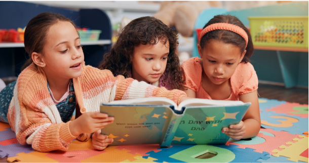 three girls lying on a playroom floor reading a children’s book together