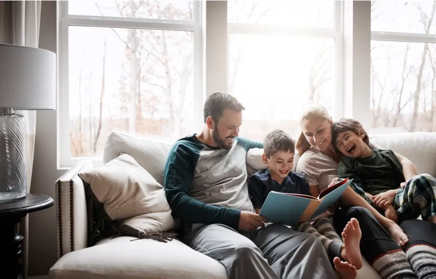 parents and two boys sitting on a couch, laughing and reading a book together