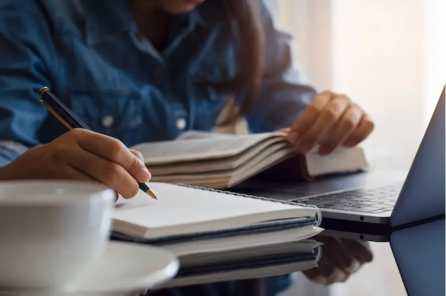 close-up of a woman writing in a notebook with an open book and a laptop in front of her