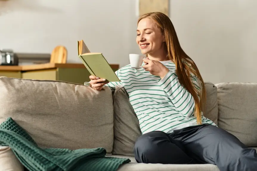 woman sitting relaxed and smiling on a couch with a book in one hand and an espresso in the other