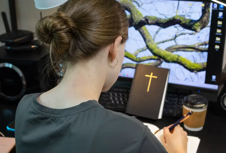 woman writing at her desk with a bible propped up in front of her against her computer screen